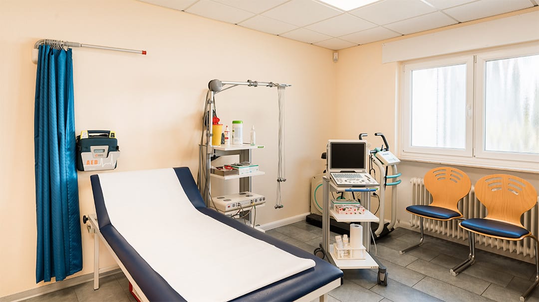 Behandlungsraum einer Arztpraxis mit Untersuchungsliege, Ultraschallgerät, Belastungs-EKG und einem Defibrillator (AED) an der Wand.