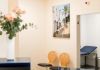 Wartebereich einer Arztpraxis vor Zimmer 4 mit zwei Holzstühlen, einem Wandbild mit Palmenmotiv und einem angedeuteten Behandlungsraum.
